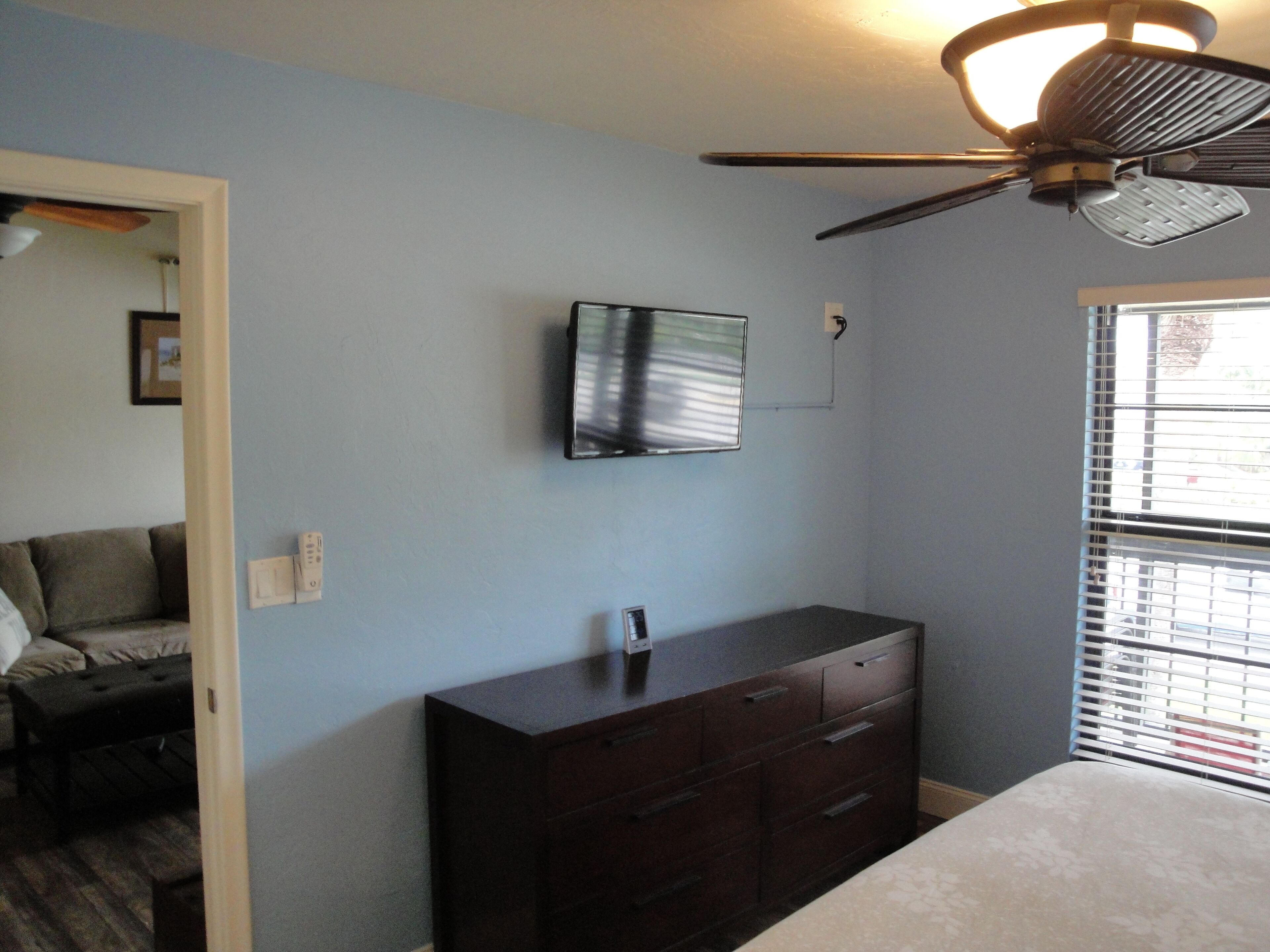 Une chambre à coucher comprenant un lit, une commode, un ventilateur de plafond, un téléviseur fixé au mur et une fenêtre avec des stores.