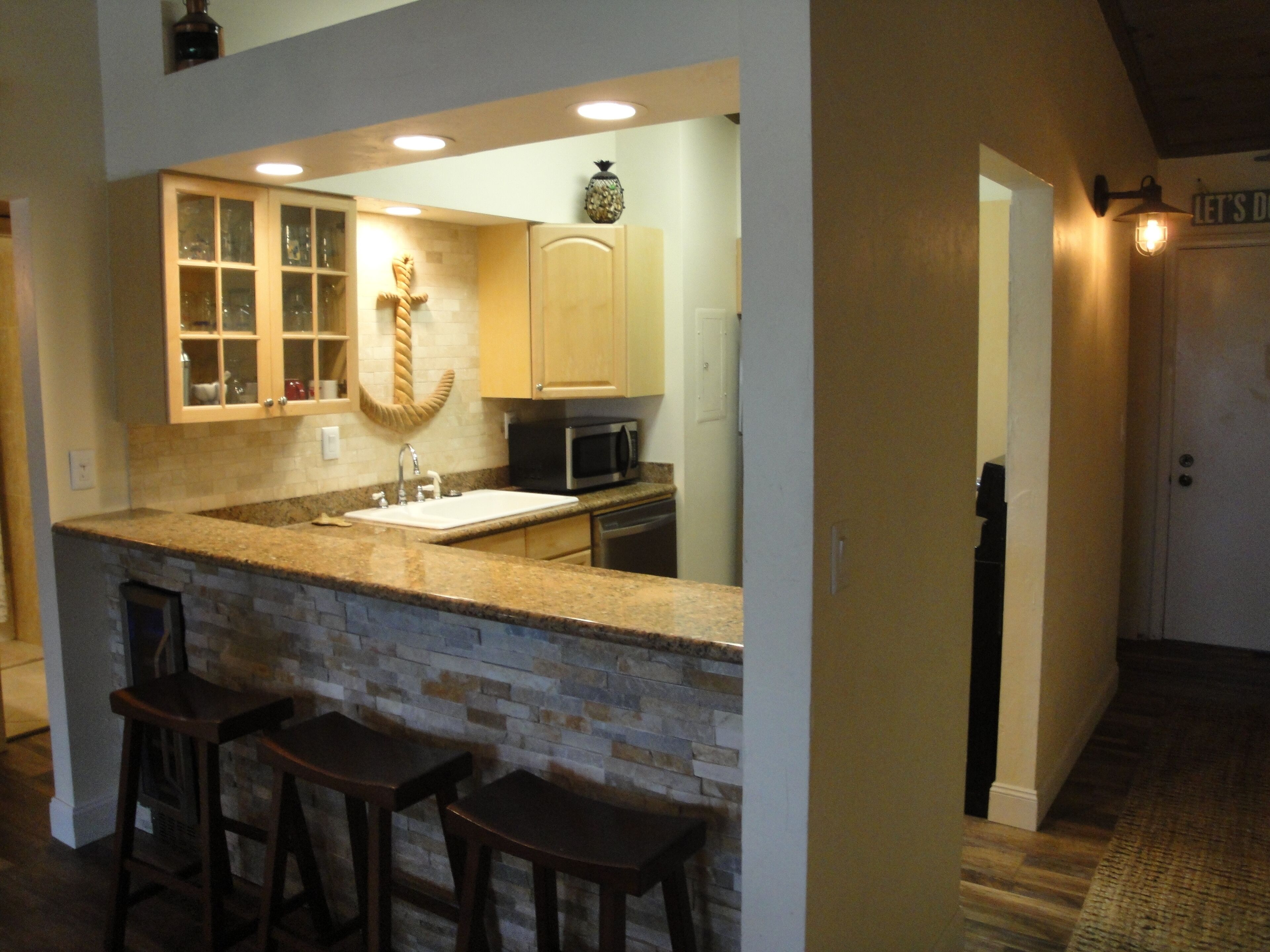 A kitchen with a granite countertop, a built-in sink, and a stone backsplash. There are three wooden bar stools and a decorative pineapple on the wall.