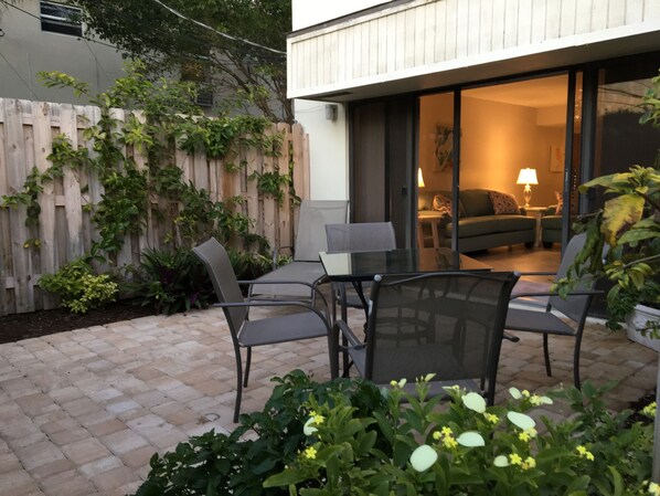 An outdoor patio with a glass door leading to a living room, surrounded by a wooden fence and greenery.