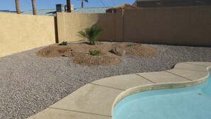 Piscina com borda de pedras e cascalho, uma pequena palmeira e um muro de concreto.
