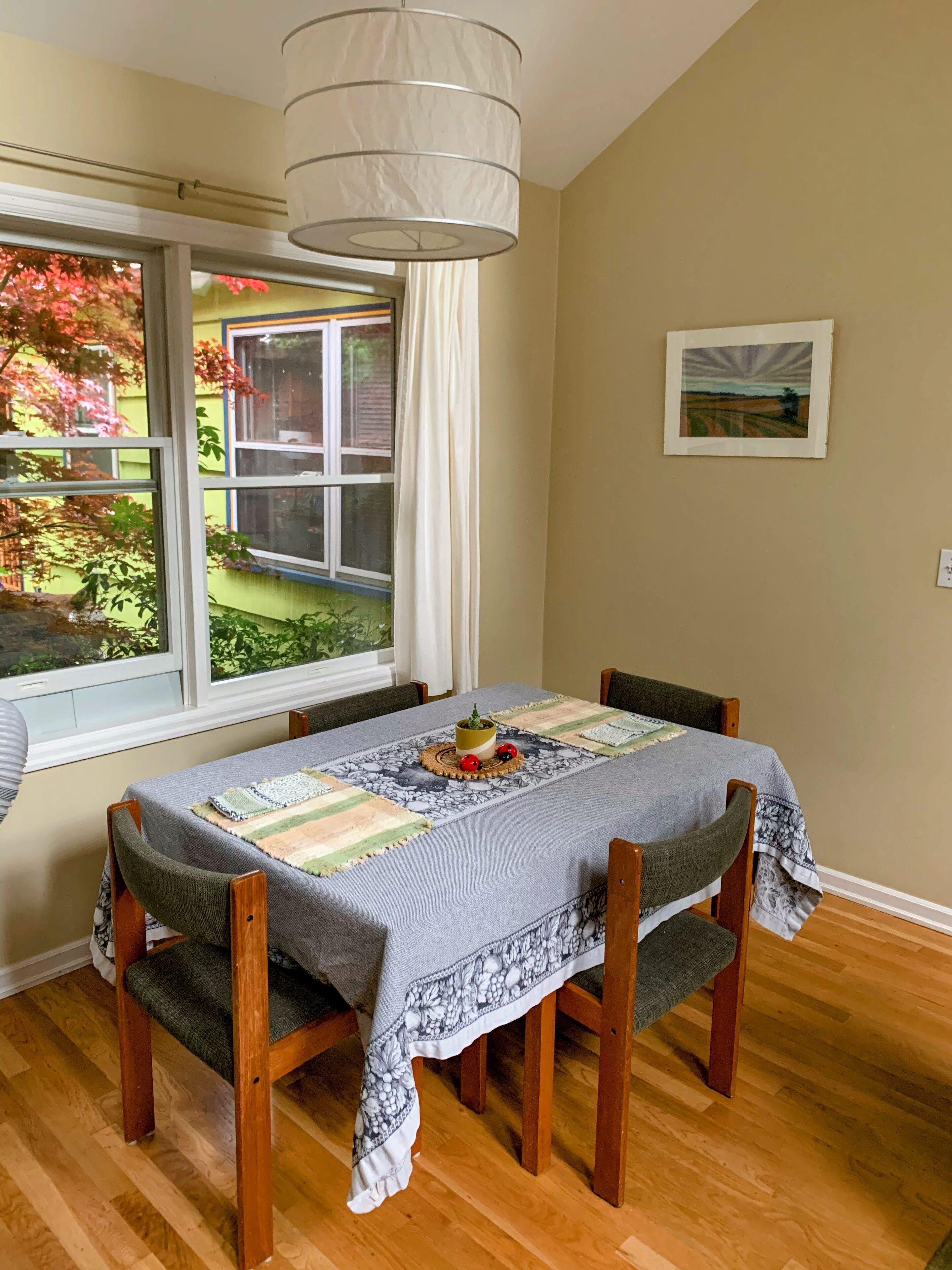 A dining area with a table set for four, a hanging light fixture, a window with a view of a garden, and a framed picture on the wall.