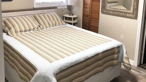 Quarto com cama, luminária de cabeceira, janela com persianas, espelho e pintura na parede.