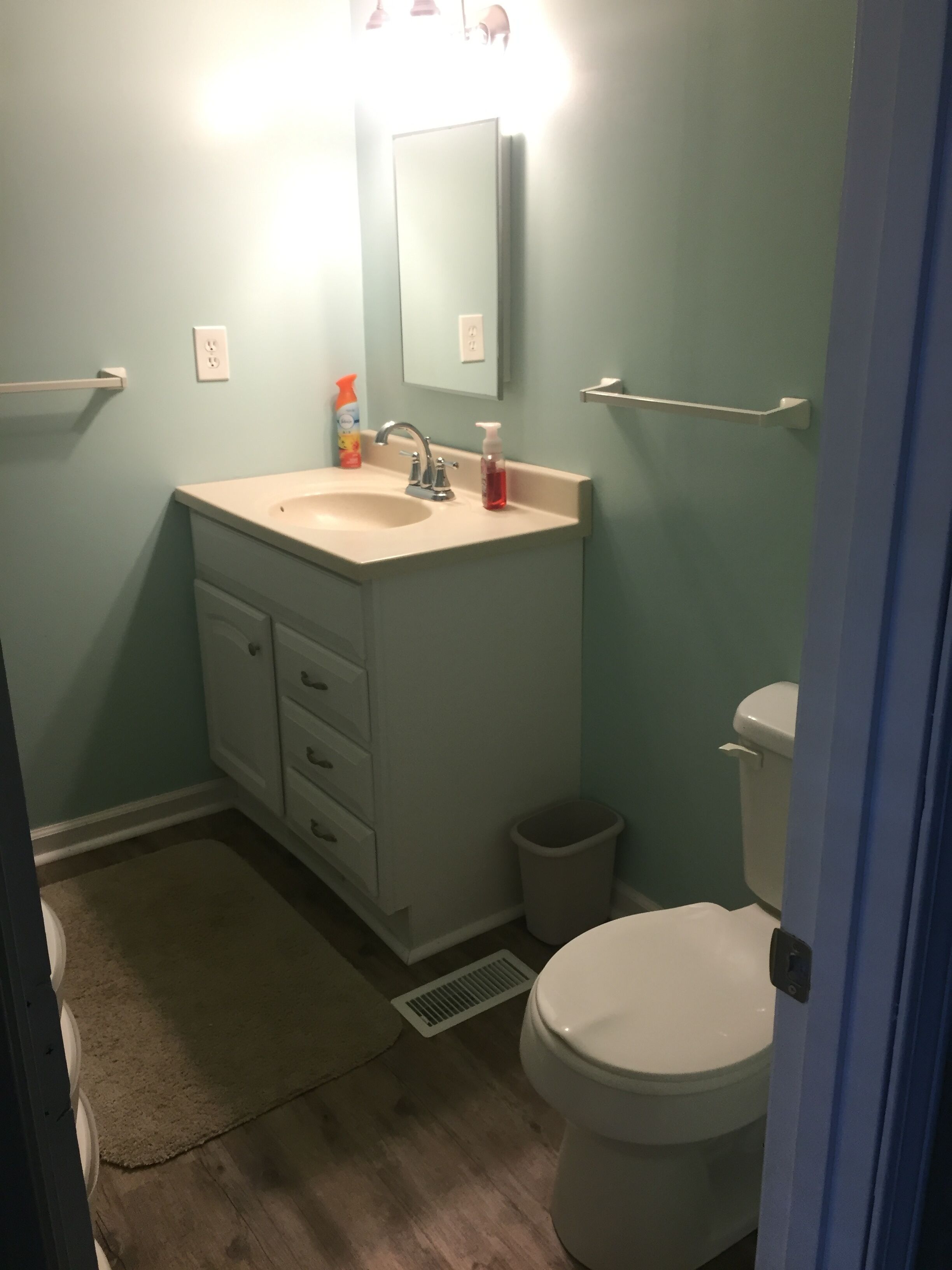 A bathroom with a sink, mirror, and toilet. There is a trash can and a towel rack.