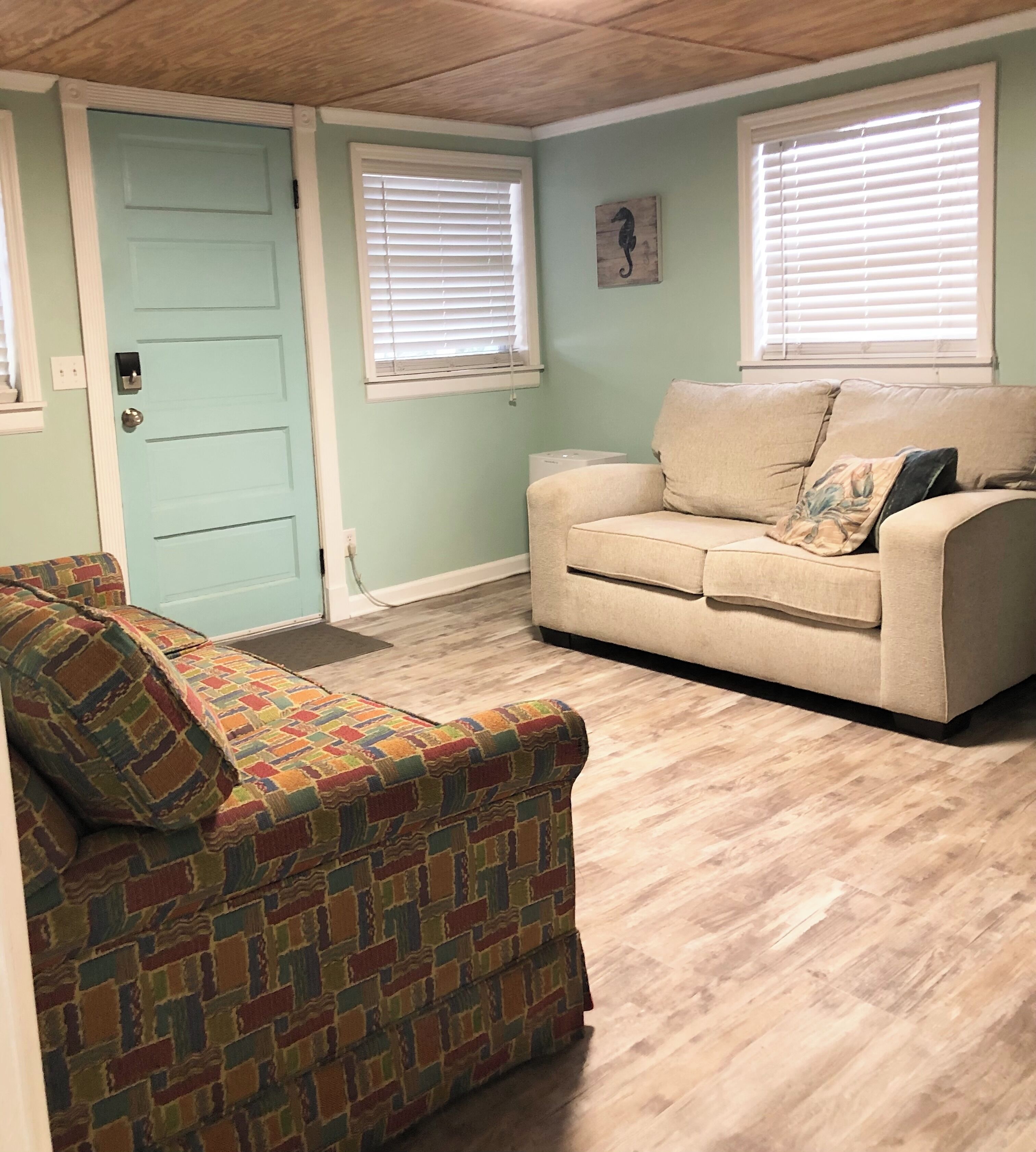 A living room with a light blue door, beige sofa, and a colorful patterned ottoman.