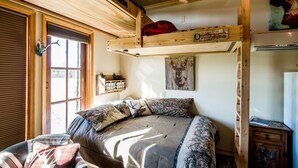 A cozy room with a bed, a wooden bunk bed above, a window with blinds, and a welcome sign.
