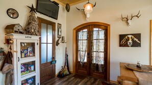 A rustic interior with a wooden ceiling, a television mounted on the wall, a deer skull, and a hunting rifle leaning against the door.