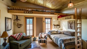 A cozy room with a loft bed, a sofa, and a chair. There is a ladder on the right side and a wooden ceiling.