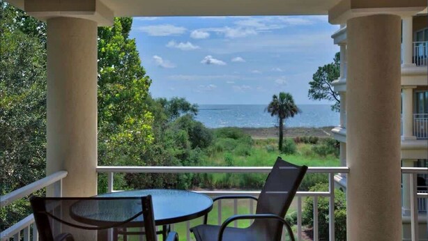 A balcony with a table and chairs, overlooking a coastal landscape with trees and a view of the sea.