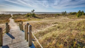 A wooden boardwalk leading to a beach with sparse vegetation and a clear sky.
