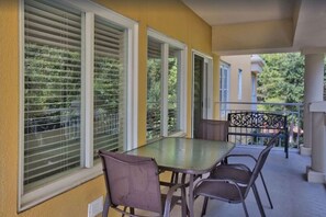 Un balcón con mesa y sillas, ventanas amplias con persianas y vistas al verdor.