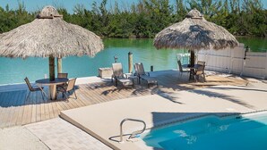 Een zwembadterras met rieten parasols, ligstoelen en een houten terras met uitzicht op een waterpartij.
