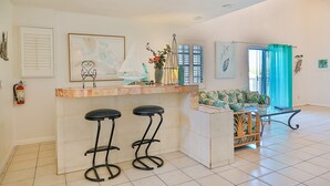 Een ruime keuken met een marmeren aanrecht, twee zwarte barkrukken, een klein eiland en een eethoek met een rieten tafel en stoelen.