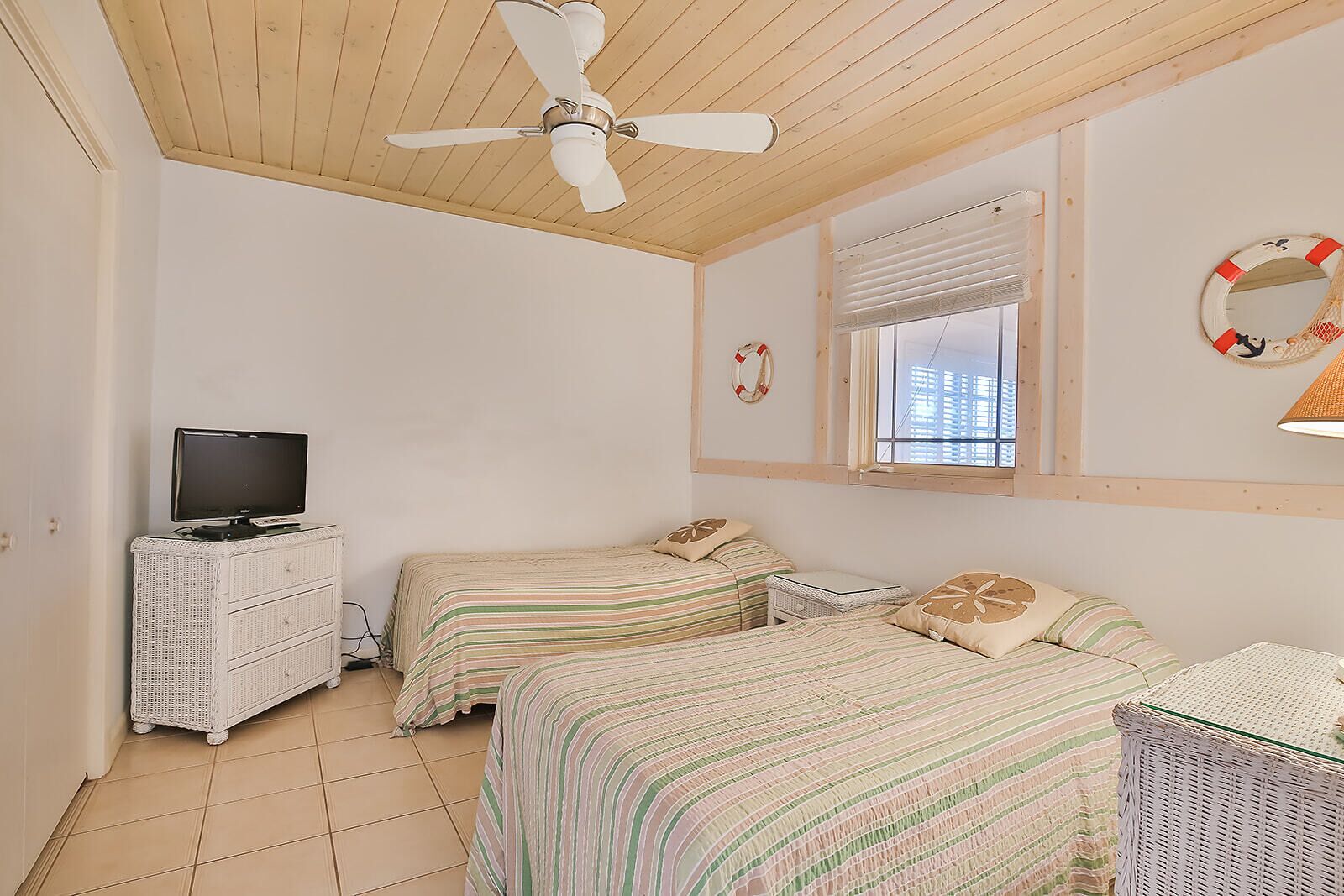 Un dormitorio con dos camas, un ventilador de techo, un televisor sobre un tocador y una ventana con persianas.