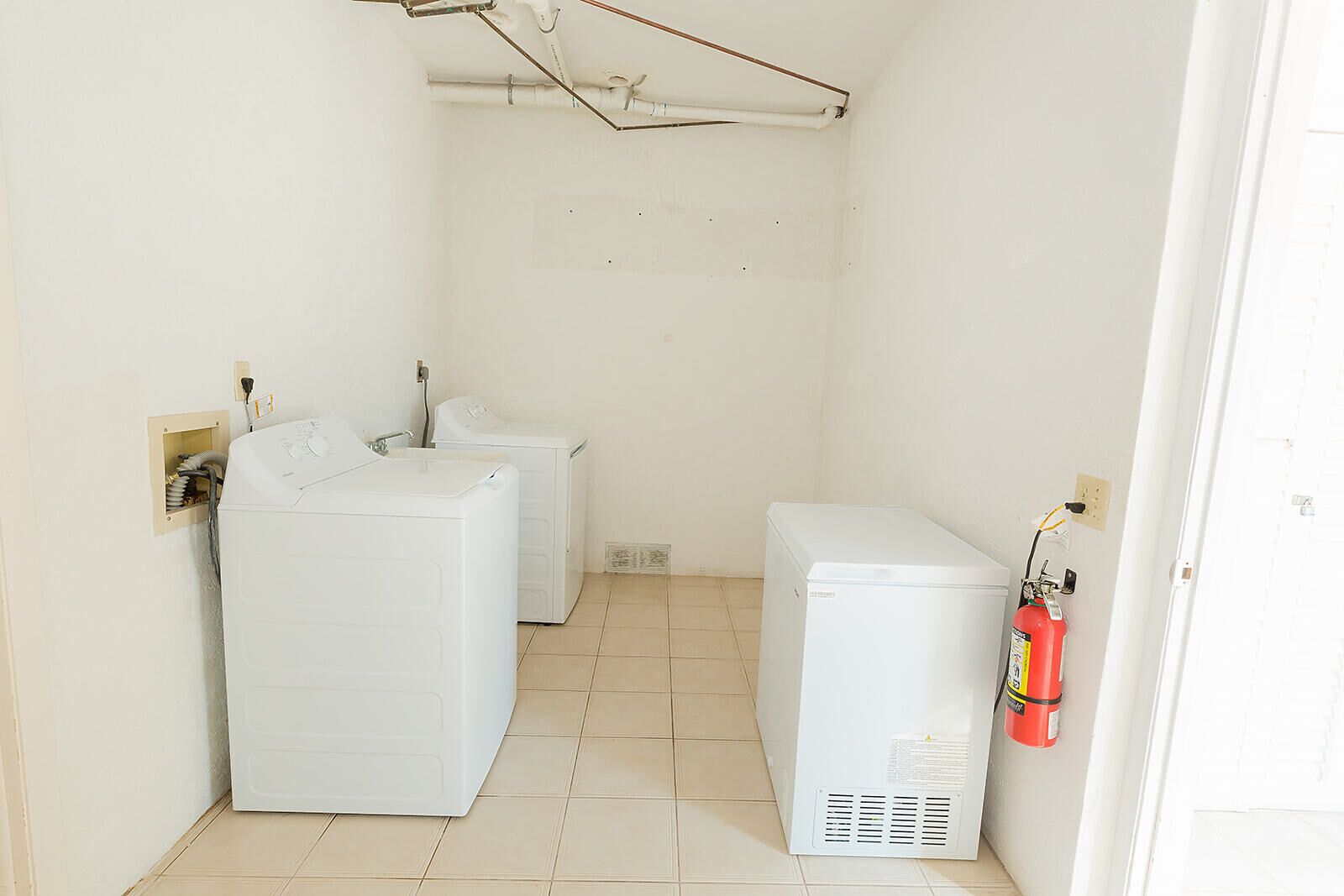 A laundry room with two washing machines and a dryer, a fire extinguisher on the wall, and exposed pipes on the ceiling.