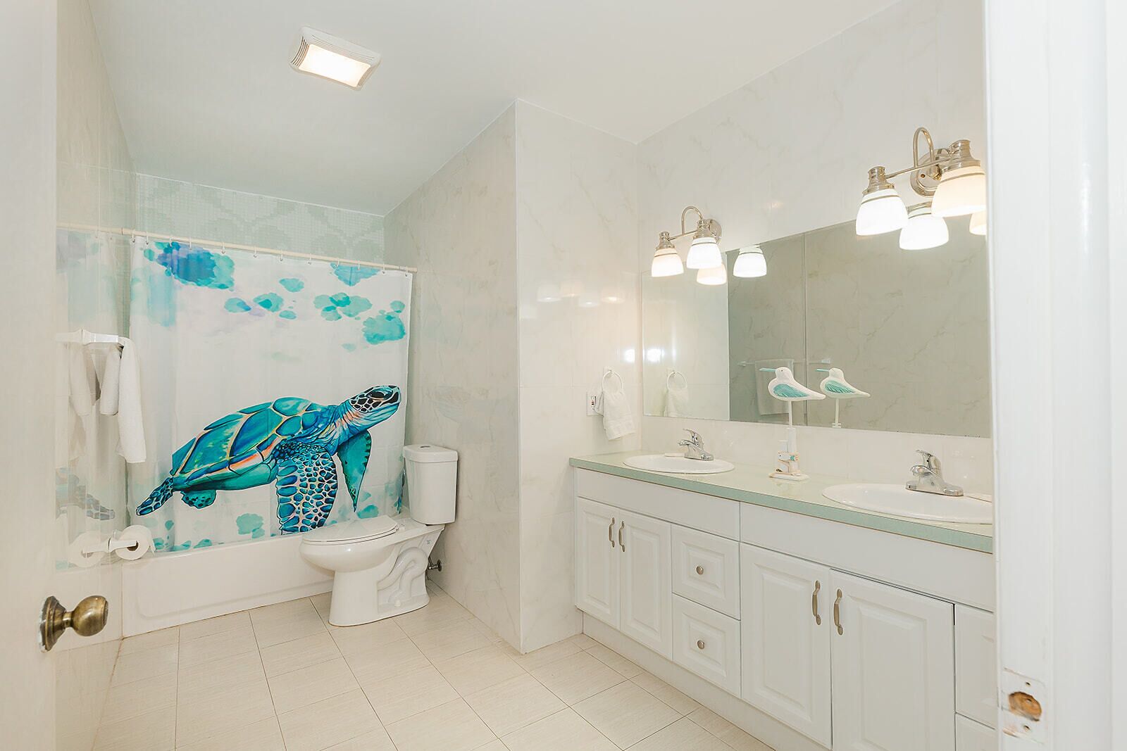 Une salle de bain dotée d’un grand miroir, de deux lavabos, de toilettes et d’une douche ornée d’une fresque représentant une tortue de mer.