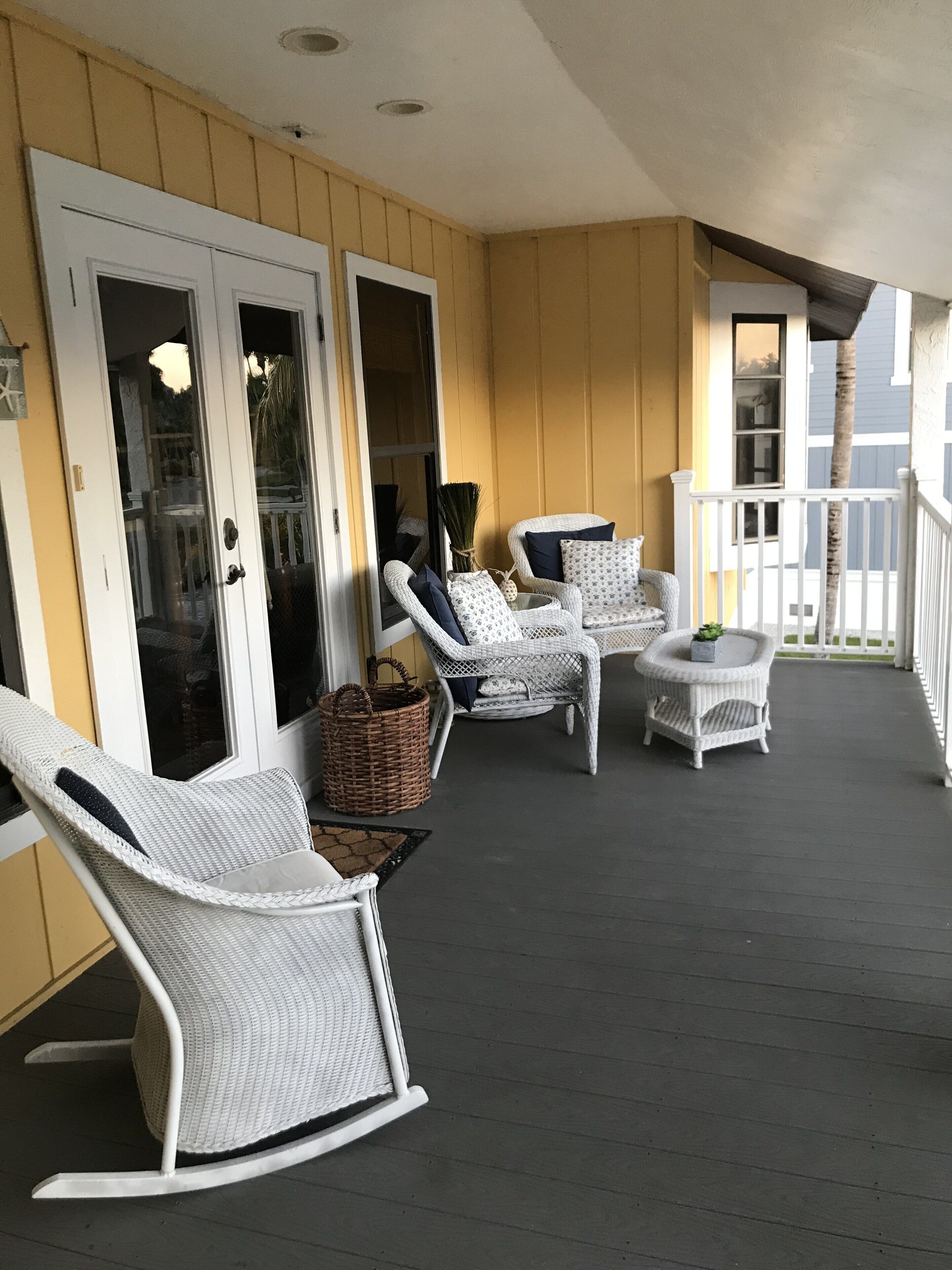 A covered porch with white wicker furniture, including rocking chairs and a small table, against a yellow wall.