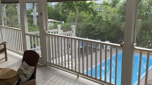 A balcony with a view of a swimming pool, white railings, wicker furniture, and a ceiling fan.
