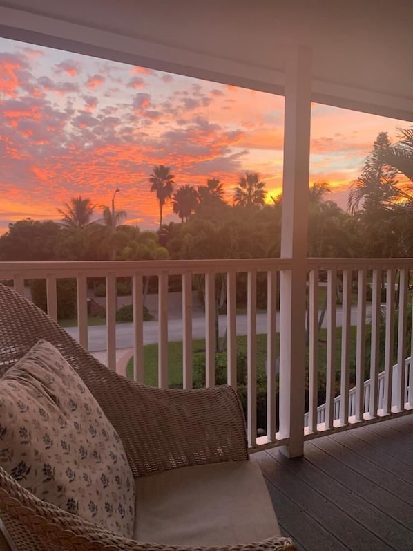 A covered porch with a wicker sofa and a view of a sunset over palm trees and a road.