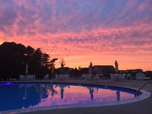 Una piscina con sillas de descanso y sombrillas, rodeada de árboles y edificios, durante el atardecer.