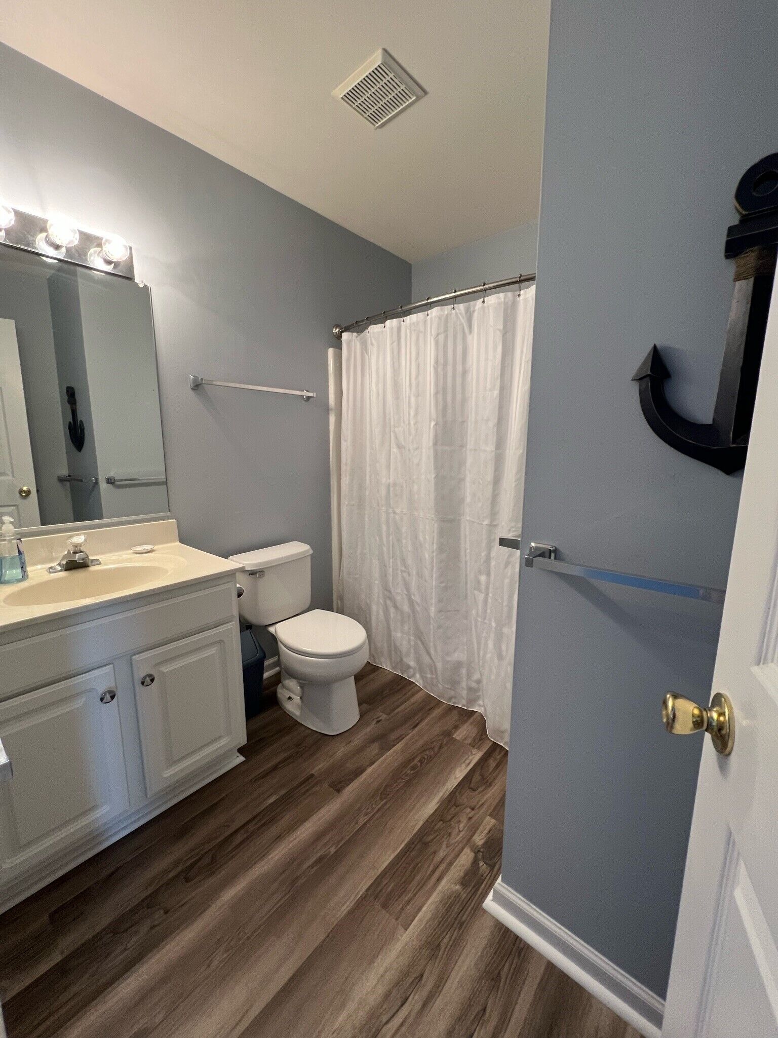 A bathroom with a wooden floor, white fixtures, a shower curtain, and a mirror with lights.