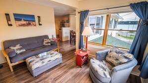 Una sala de estar acogedora con un sofá, un sillón y una mesita con una lámpara. Una ventana ofrece vistas a un lago y edificios.