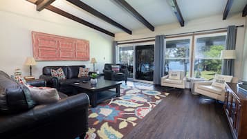 A living room with a dark wood floor, a patterned rug, a black leather sofa, a coffee table, and a large window with curtains.