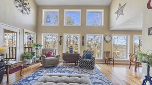 A spacious living room with multiple windows, a large tufted ottoman, and various seating options. - Hartwell Lake deep water home, family friendly and close to Clemson and UGA (Lavonia)