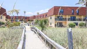 A pathway leading to a row of yellow buildings with red roofs, surrounded by tall grasses and palm trees.