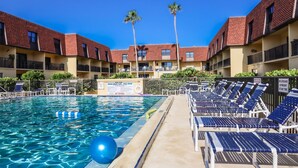 A swimming pool with lounge chairs and palm trees, surrounded by a building with red-tiled roofs.