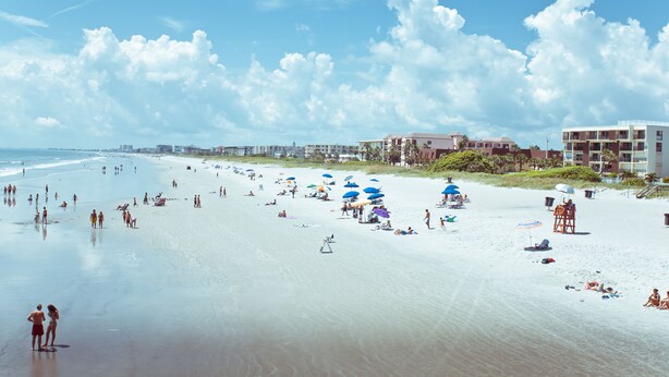 A beach with umbrellas, people, and buildings in the background.