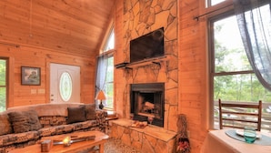 A cozy cabin interior with a stone fireplace, wooden walls, a patterned sofa, and a dining area with a table and chairs. - Beautiful, pet friendly secluded cabin with fire place and hot tub. (Robbinsville)