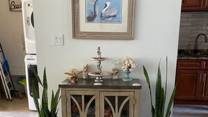 Un salon avec un meuble en bois, deux plantes en pot, un tableau encadré et, au fond, un coin cuisine.