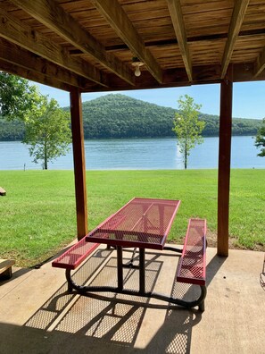 Un espace pique-nique abrité, équipé d’une table et de bancs en métal, offrant une vue sur un lac et des collines boisées.