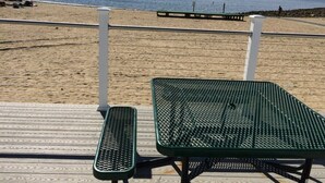 Eine Strandterrasse mit einem grünen Metalltisch und einer Bank, mit Blick auf einen Sandstrand und ruhiges Wasser mit Booten.