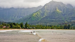 A beach with people swimming and surfing, a mountain range in the background, and wind turbines on the mountains. - Newly Remodeled Ocean View Condo, 2 bedroom, 2 bath, No stairs!  (Princeville)