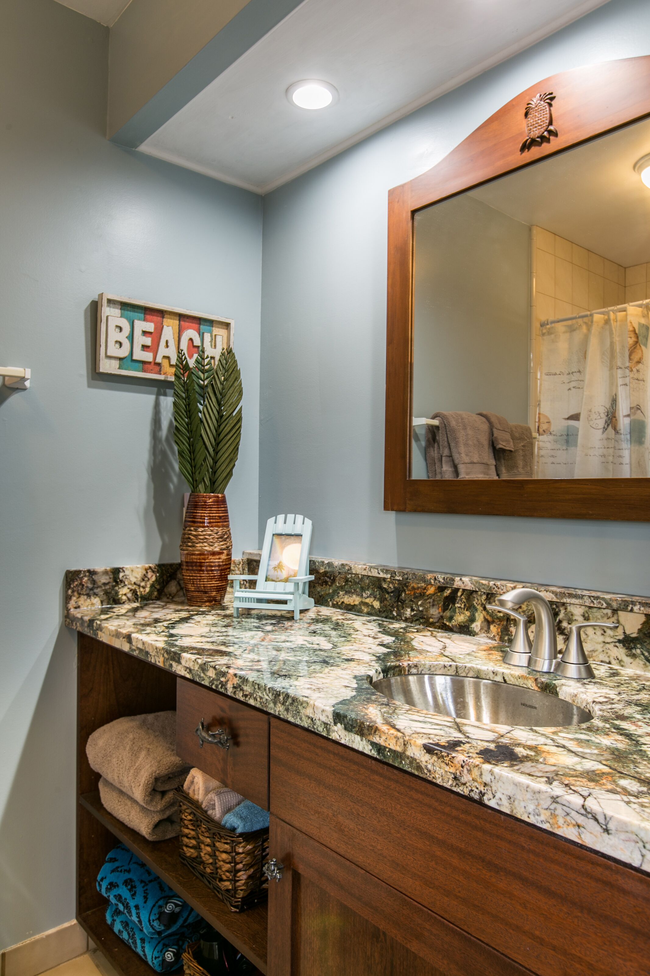 A bathroom with a marble countertop, wooden vanity, a mirror, a towel rack, and a wall-mounted sign that reads "BEACH".
