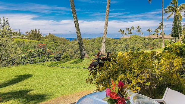 A round glass table with a flower vase, surrounded by greenery and palm trees, with a clear view of the ocean in the background. - Newly Remodeled Ocean View Condo, 2 bedroom, 2 bath, No stairs!  (Princeville)