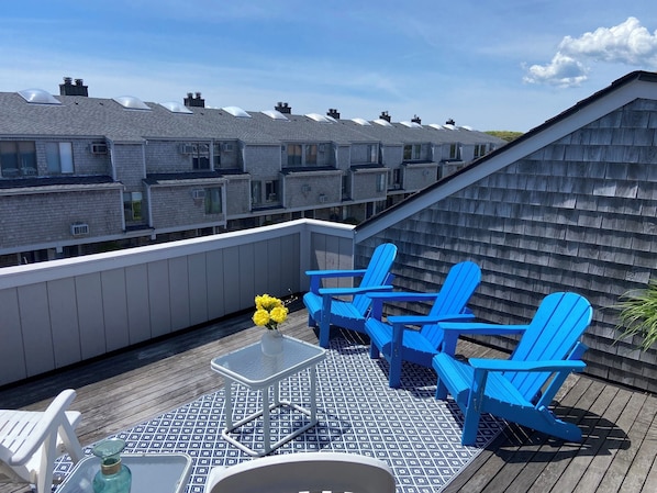 A rooftop deck with blue Adirondack chairs, a small table, and a vase of yellow flowers.