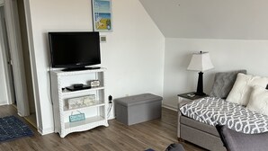 A modern bedroom with a flat-screen TV, a bed with patterned bedding, a side table with a lamp, and a small storage unit.