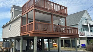 A two-story wooden deck structure with railings, supported by wooden posts, attached to a house with a gabled roof and white siding.