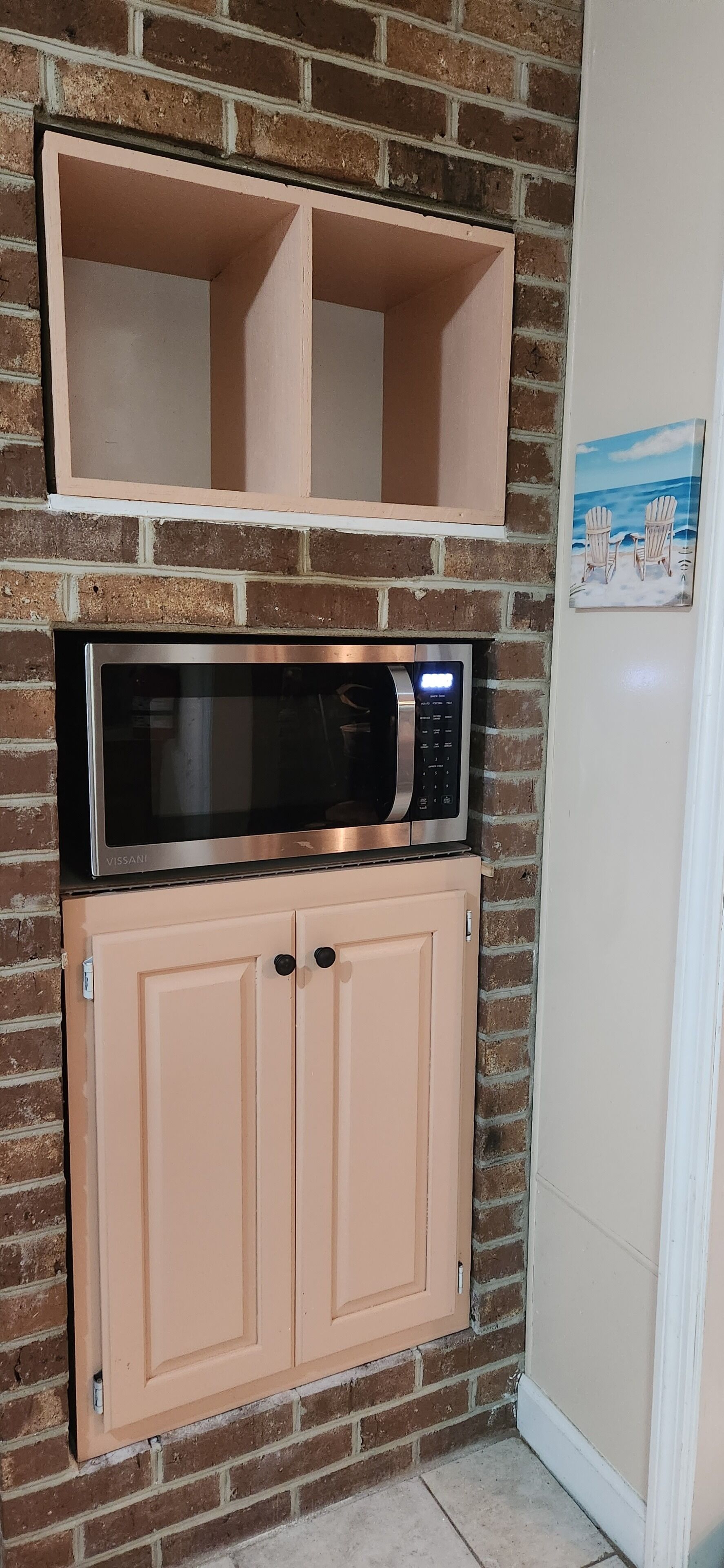 A kitchenette with a VISSANI microwave above a cream-colored cabinet, a brick wall, and a beach-themed picture on the adjacent wall.
