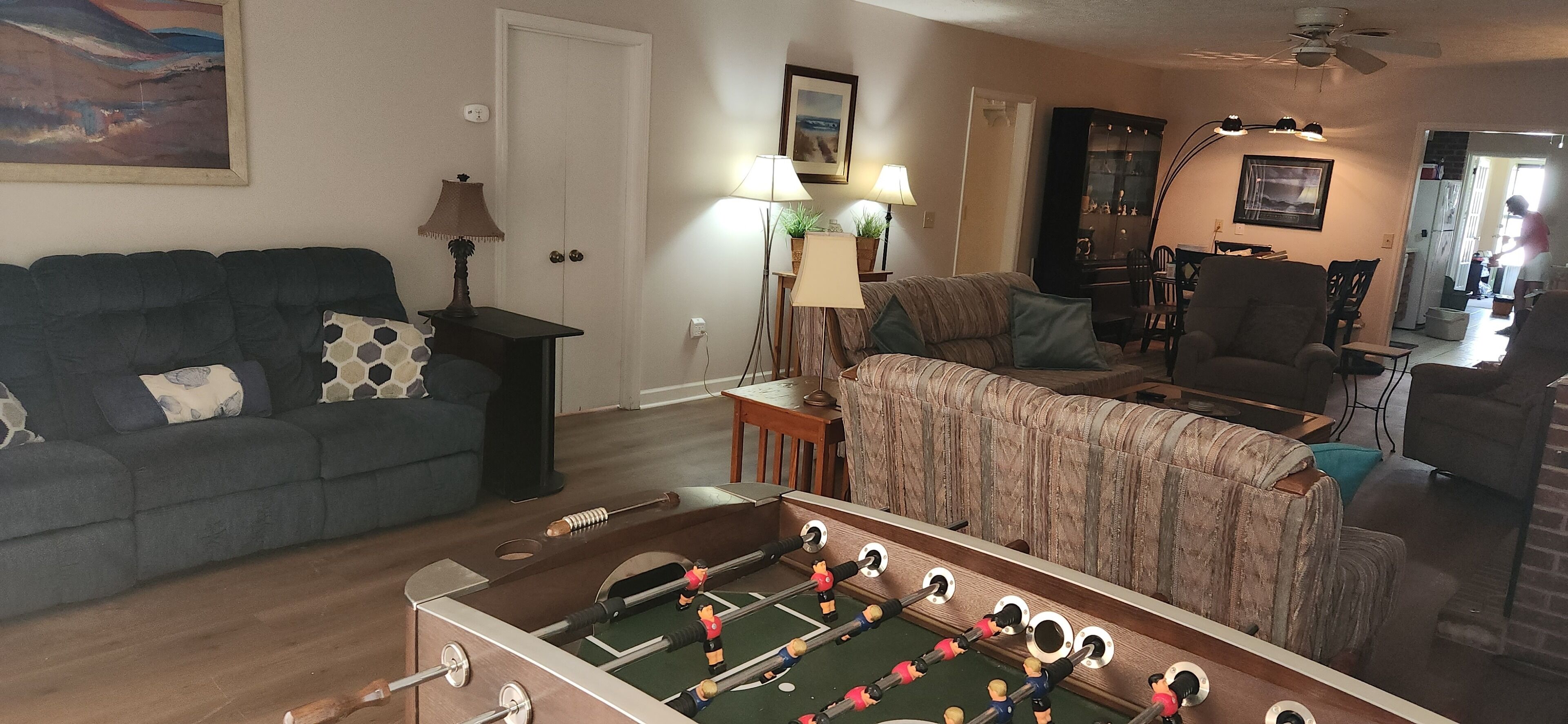 A living room with a foosball table, a sofa, a dining table, and a kitchen area in the background.