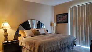 A bedroom with a large bed, two bedside tables with lamps, a painting on the wall, and a window with blinds. - 1-Level Patio Home Great Location 1 Block from Ocean - NO STEPS! w/ 1-car garage (North Myrtle Beach)