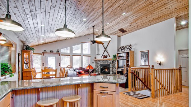 A spacious kitchen with wooden accents, a large island with stools, a stone fireplace, and a wooden staircase. - Serenity Pines (Yosemite National Park)