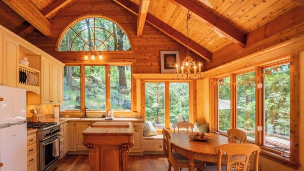 Interior de una cabaña de madera con cocina, comedor y ventanas amplias que dan a un bosque.