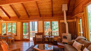 Interior acogedor de una cabaña con paredes y suelos de madera, una zona de comedor, una sala de estar con sofá y chimenea, y ventanas amplias con vistas al bosque circundante.
