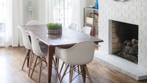 Un comedor con una mesa de madera, sillas blancas, una lámpara colgante y una chimenea con pared de ladrillo blanco.