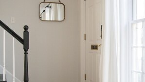 Un pasillo con piso de madera, una puerta blanca, un espejo en la pared y una escalera con barandilla negra.