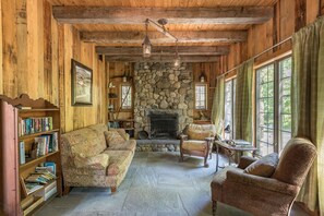 A cozy cabin interior with a stone fireplace, wooden walls, a sofa, a chair, and a bookshelf.