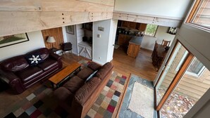 Een ruime woonkamer met een hoge plafond, houten plafond, een bruine leren bank, een houten salontafel, en een keuken met houten kasten en een eettafel.